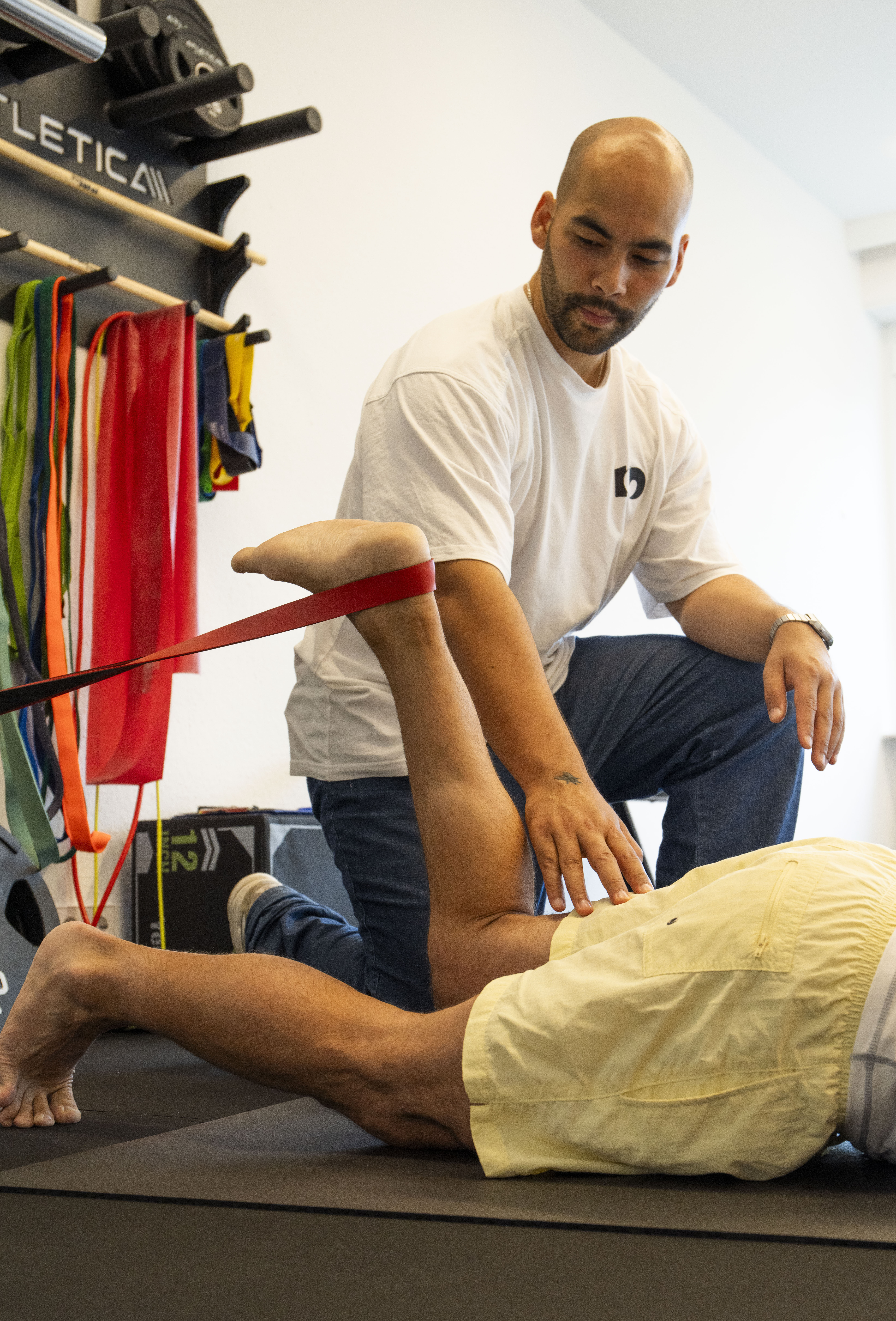 Physiotherapeut korrigiert Patient bei einer Übung in Bauchlage. Patient hat ein Gymnastikband um das Sprunggelenk und zieht die Ferse in Richtung Gesäß.